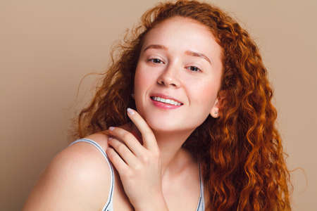 A beautiful red-haired girl with long curly hair and small freckles looks at the camera and smiles. Studio shot on a beige background. The concept of natural beauty.の写真素材