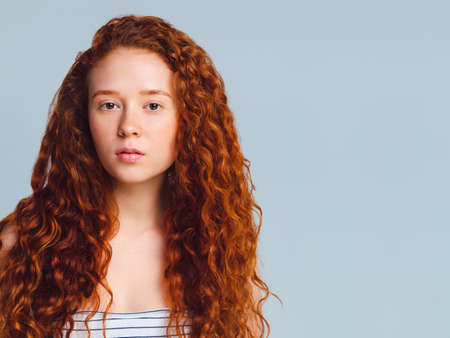 pretty teenage girl with luxurious long curly red hair on a blue background in the studioの写真素材