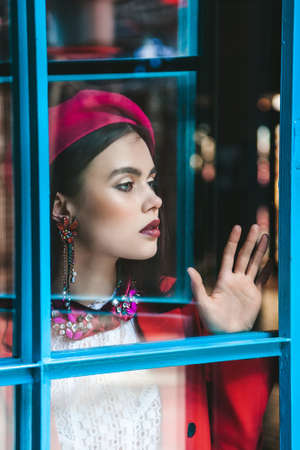 very beautiful brunette model with blue eyes in a red beret is looking through the glass. Skin retouching.の写真素材