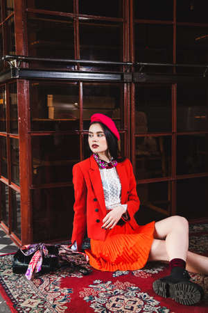 young beautiful brunette girl in a red suit, white shirt and red beret sits on the floor and looks awayの写真素材