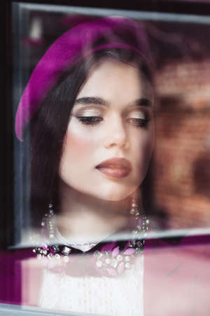 a beautiful girl in a purple jacket and beret is standing behind the glass, a reflection of the window frame on her face. Close-up portraitの写真素材