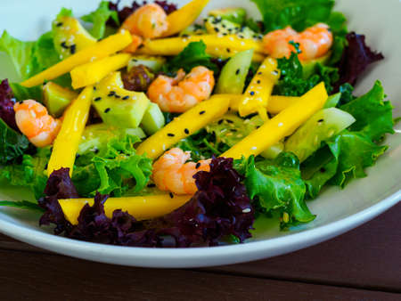 appetizing salad of shrimp, mango, avocado, cucumber, mix of lettuce leaves, decorated with black sesame seeds, on a white plate, close-upの写真素材