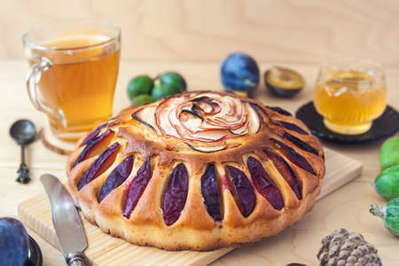 round cake decorated with slices of plums and apples. Autumn still life with fresh fruits and tea on a light wooden tableの写真素材