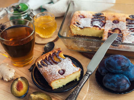 a square slice of plum pie on a round black antique plate, acacia honey, a vintage teaspoon and a cup of black teaの写真素材