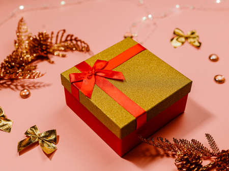 gift red box with gold lid tied with satin ribbon, shiny sprigs with cones, bows, beads and garland on a peach backgroundの写真素材
