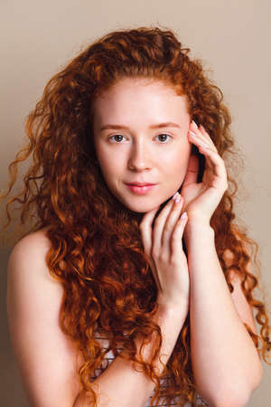 Portrait of happy girl smiling on camera on beige background. Model with long natural red curly hair and perfect skin with little freckles without cosmetics. Young skin care conceptの写真素材