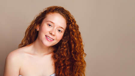 beautiful girl with thick red long curly hair smiles in the studio on a beige background. Copy spaceの写真素材