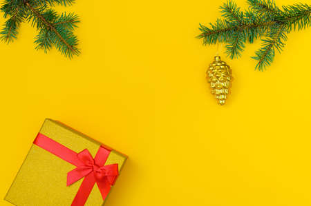 horizontal festive yellow background: shiny gift box with red ribbon, fir branches and Christmas tree toy - golden cone. Copy space centerの写真素材