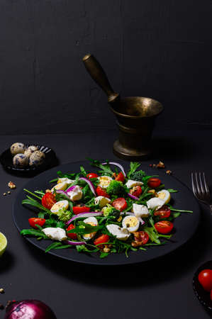 diet salad of arugula, quail eggs, cherry tomatoes, broccoli, purple onions, cream cheese and walnuts on a black matte plate on a dark background. Still life of dish, ingredients and antique copper mortarの写真素材