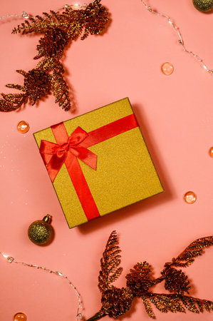 gift red box with gold lid tied with satin ribbon, shiny sprigs with cones, bows, beads and garland on a pink backgroundの写真素材