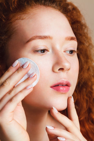 Young skin care concept. Attractive teenage girl with long red curly hair rubs her face with a cotton pad and looks at the camera. No makeup. Natural beauty.の写真素材