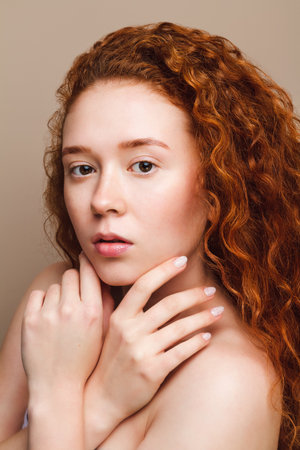 young beautiful girl with long red curly hair looks at the camera. The concept of natural beauty. Close-up shot in the studio on a beige background, verticalの写真素材