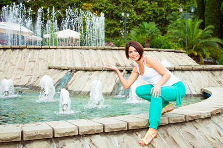 beautiful young brown-haired woman in a white T-shirt and turquoise pants near the fountain. Model smiling and looking at the cameraの写真素材