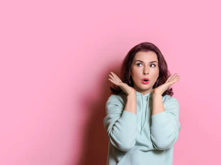 surprised young woman in light green hoodie looks at copy space. Horizontal studio shot on pink backgroundの写真素材