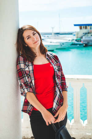 a beautiful brunette in a red T-shirt stands near a column in the port against the backdrop of yachts and boats. The model looks into the camera and smiles. Vacation and travel conceptの写真素材