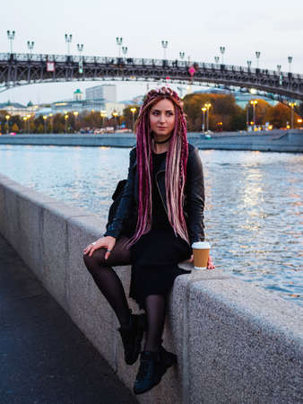 beautiful Caucasse woman with long pink dreadlocks and pigtails in black clothes sits on the parapet of the Moscow River embankment in the eveningの写真素材
