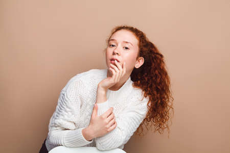 beautiful girl with long red curly hair in a white sweater in the studio on a beige background looks at the cameraの写真素材