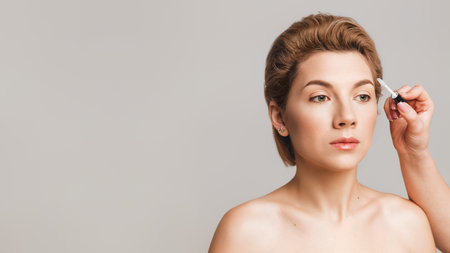 makeup artist applies eyebrow gel with a brush. Beautiful caucasian model with green eyes on a gray background. Make-up application concept. Copy spaceの写真素材