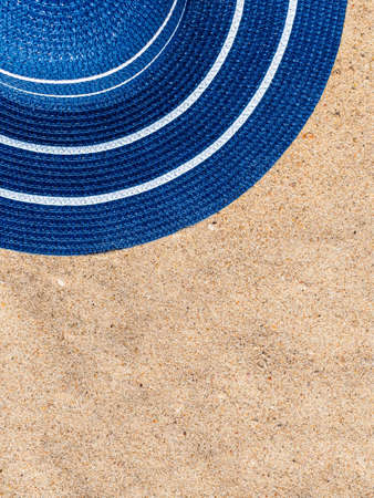 Vertical background: a blue straw hat with white stripes lies on the sand on the beach, copy space for your text. Vacation concept by the seaの写真素材