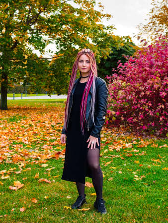 beautiful young woman with long pink dreadlocks in black dress, biker jacket and sneakers posing in the park on the grass in autumnの写真素材