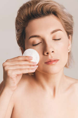 beautiful model - a blonde with short hair removes makeup with a cotton pad. The concept of skin cleansing. Studio shot, close up, retouchingの写真素材