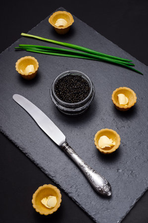 natural black sturgeon caviar in a glass jar, vintage knife, shortcrust pastry tartlets with butter, green onion feathers on a slate plate close-upの写真素材