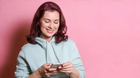 smiling young woman in mint hoodie looks at the smartphone screen. Social media communication concept. Studio shot over pink background, copy space.の写真素材