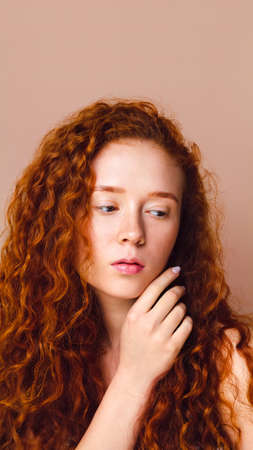 portrait of red-haired girl with long curly hair and freckles on beige background in the studio. Skin care concept. Copy spaceの写真素材