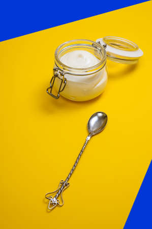 thick greek yogurt in glass jar and vintage dessert spoon on yellow and blue backgroundの写真素材