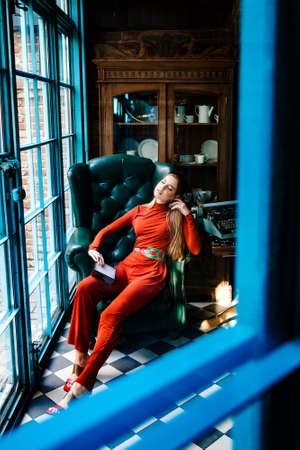 attractive tall young woman with long blond hair in a red jumpsuit and headband sits in a chair in a...