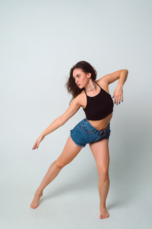 young beautiful woman with brown hair in a black top and short denim shorts dances barefoot on a white background. full length portraitの写真素材