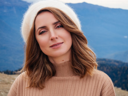 close-up portrait of a beautiful blonde woman in a beige beret and sweater against the backdrop of mountains in springの写真素材