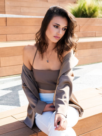 very beautiful brown-haired woman in a beige satin jacket and a tank top sits on a wooden bench in the parkの写真素材