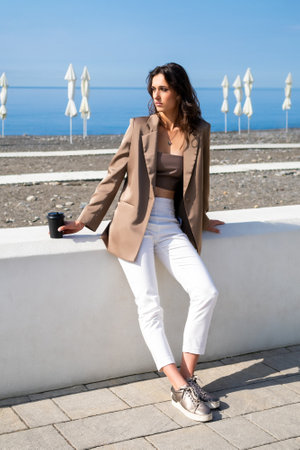tall, slender brunette is sitting on the sea embankment and looking away. A full-length portrait of a beautiful woman in a brown jacket, top and white trousers against the background of the sea. Fashion model posing in the harsh sunlightの写真素材
