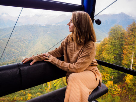 an attractive blonde model sits in a cable car cabin and admires the view. Beautiful woman climbs the funicular in the mountains in autumnの写真素材