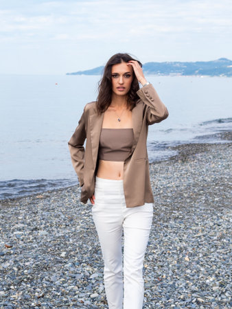 beautiful slender brunette in a beige jacket, tank top and white trousers walks along the beach in autumnの写真素材