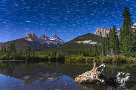 Star trails above The Three Sisters trio peaks mountain at night, starry sky reflected in the creek water surface. Beautiful landscape in Banff National Park, Canadian Rockies, Alberta, Canada.の写真素材