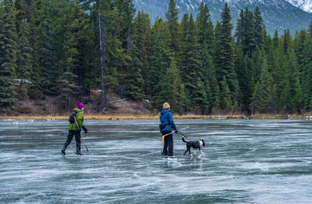 Tourists doing ice-skating in Johnson Lake frozen water surface in winter time. Snow-covered mountain in the background. Banff National Park, Canadian Rockies, Alberta, Canada.のeditorial素材