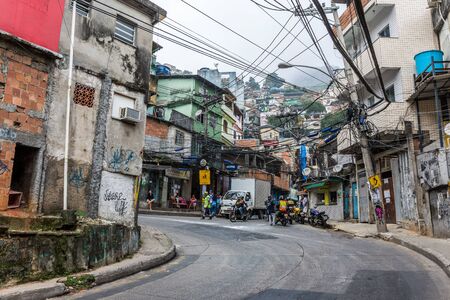 A street view looking up a road with Brazilians on scooters & cramped buildings in Rocinha favela, a...