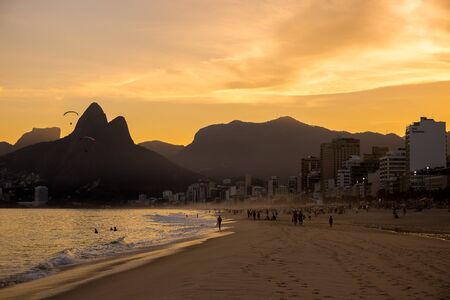 Ipanema beach with silhouette of people relaxing & having fun & paragliders overhead at sunset & Dois IrmÃ£os (Two Brothers) mountains in background in Rio de Janeiro, Brazil, South Americaのeditorial素材
