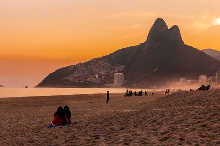 Two girls sat on Ipanema beach at sunset with people playing and relaxing & Dois IrmÃ£os (Two Brothers) mountains in background in Rio de Janeiro, Brazil, South Americaのeditorial素材