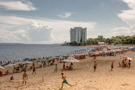 Group of local Brazilians play beach soccer while people relax sitting on chairs and swim in the black Rio Negra river at the urban Ponta Negra beach in Manaus located in the Amazon region of Brazil, South Americaのeditorial素材