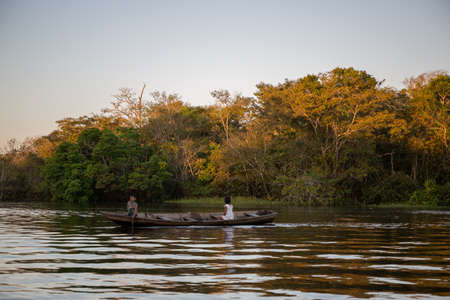 A young local Brazilian boy and girl sitting in a traditional wooden rowing boat at sunset on the Amazon river water surrounded by rainforest in the State of Amazonas in Brazil, South Americaのeditorial素材