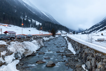 River flowing through Ischgl ski resort in Austria during winter just before nightfall with ice and snow and mountains in background with cloudy moody skyの写真素材