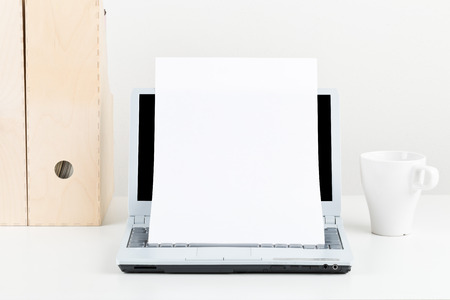 Empty sheet of paper as copy space in front of laptop on white workplace office desk with cup and...