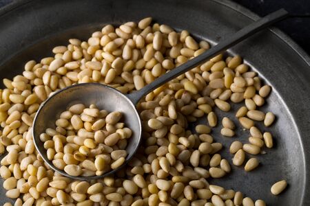 Heap of raw, uncooked pine nuts in metal spoon on rustic plate over dark kitchen backgroundの写真素材