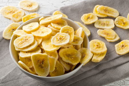 Heap of dried banana chips snack in white bowl on rustic table with grey napkinの写真素材