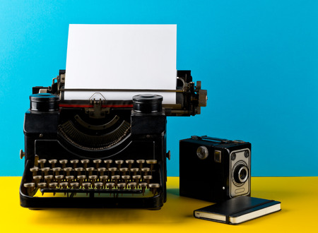 Vintage typewriter with empty, blank sheet of paper, camera and notebook on yellow and cyan...