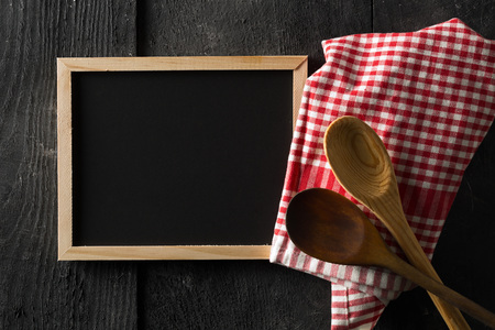 Blank, empty, black chalkboard with wooden cooking spoons and red checkered dish towel flat lay from above on black wooden table with copy spaceの写真素材
