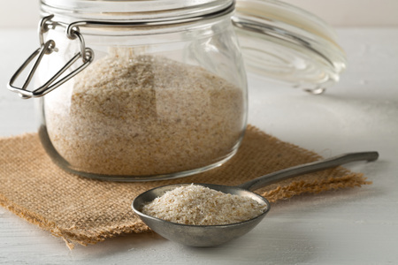 Heap of psyllium husk also called isabgol in metal spoon and glass jar on white table background. Psyllium husk also called isabgol is fiber derived from the seeds of Plantago ovata plant found in India. Selective focus.の写真素材
